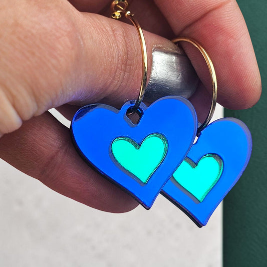 Morti's Fun Club royal blue and green acrylic heart earrings with silver-plated hoops held in a hand.