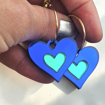 Morti's Fun Club royal blue and green acrylic heart earrings with silver-plated hoops held in a hand.