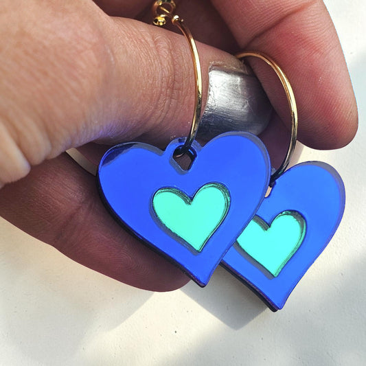 Morti's Fun Club royal blue and green acrylic heart earrings with silver-plated hoops held in a hand.