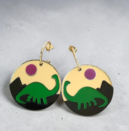 Morti's Fun Club Jurassic Moonrise circular earrings featuring a green dinosaur, black mountains, and pink mirror sun.
