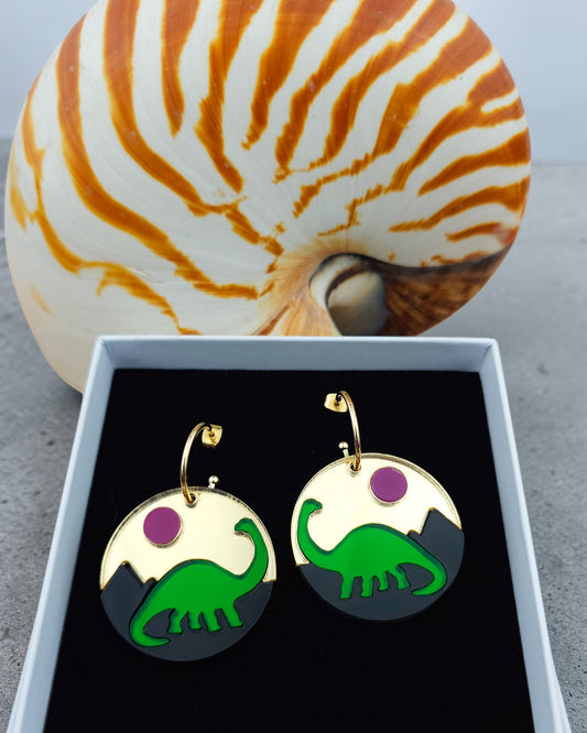 Morti's Fun Club Jurassic Moonrise circular earrings featuring a green dinosaur, black mountains, and pink sun.