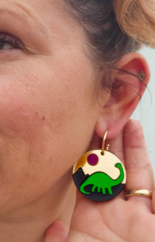 Morti's Fun Club Jurassic Moonrise earrings featuring a green dinosaur on a black and gold circular acrylic design.
