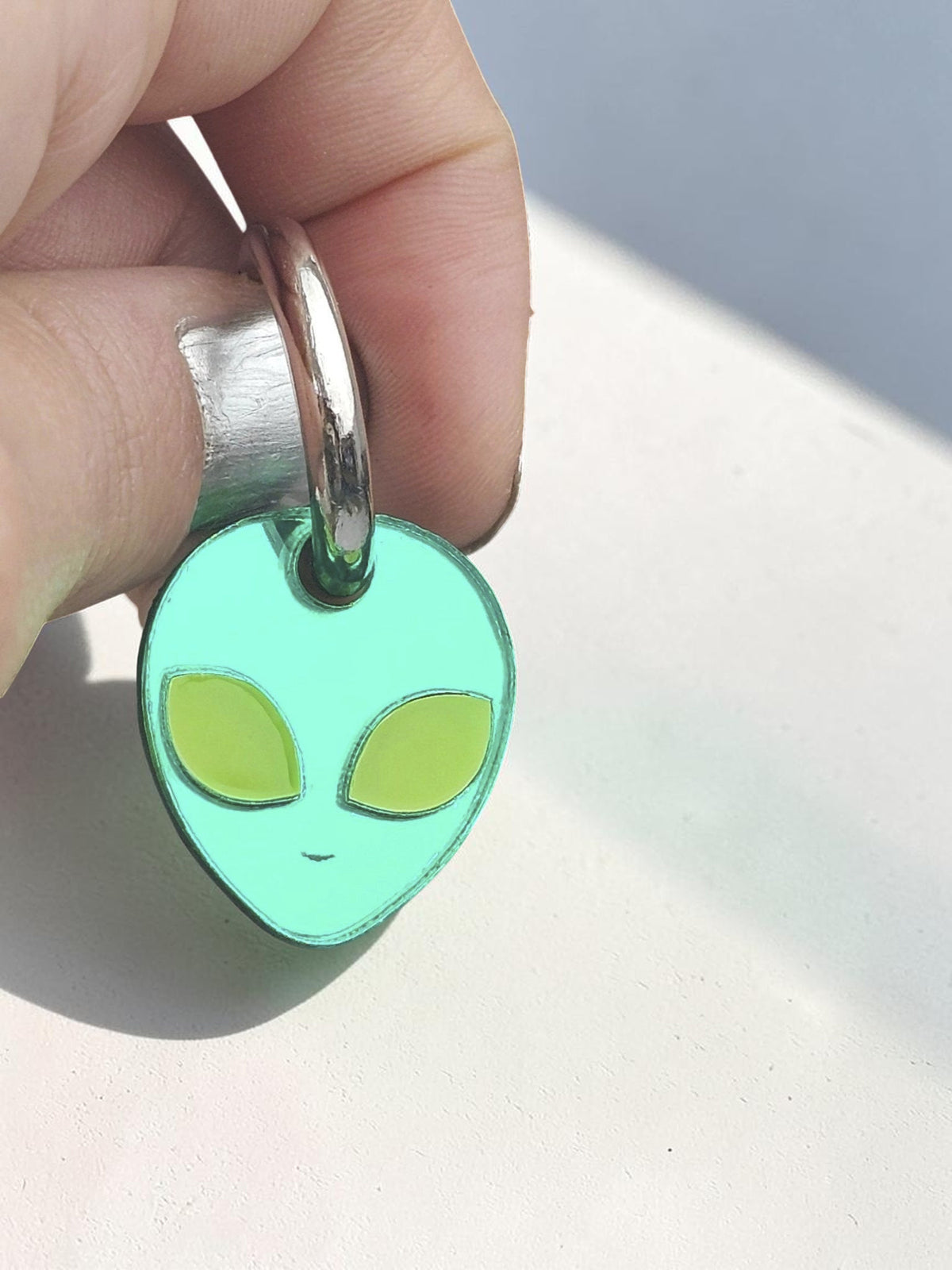Morti's Fun Club green acrylic alien charm on a silver hoop earring against a dark celestial background