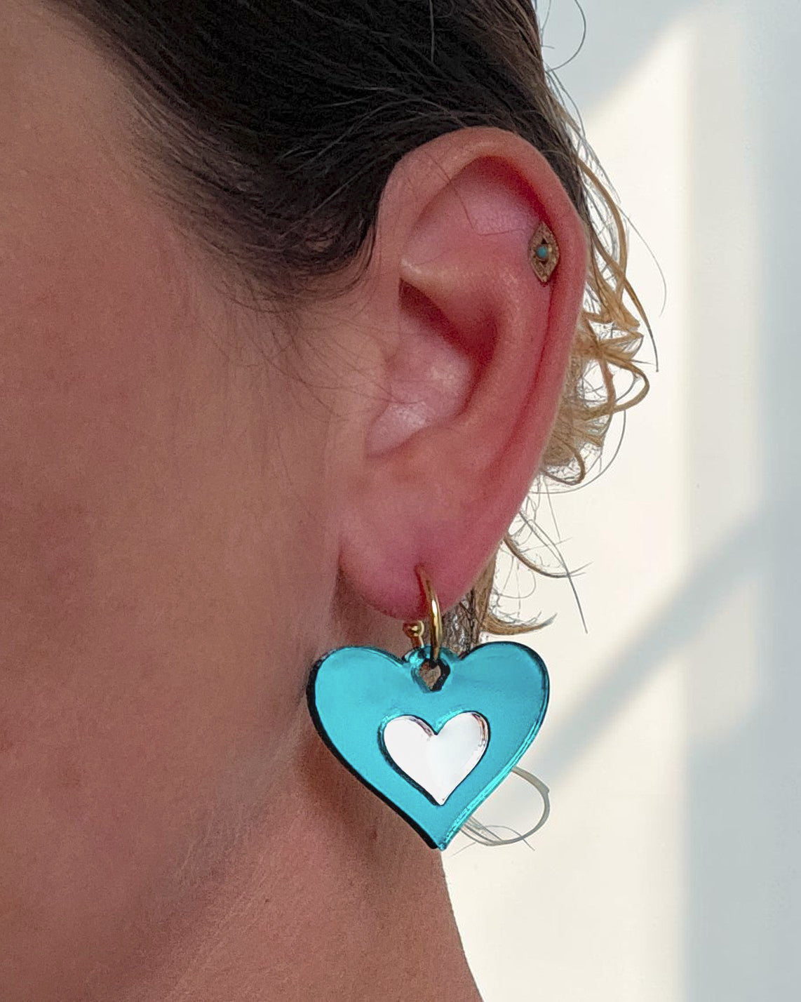 Morti's Fun Club Ice Queen heart earrings in teal and silver mirrored acrylic on a gold hoop worn on an earlobe.