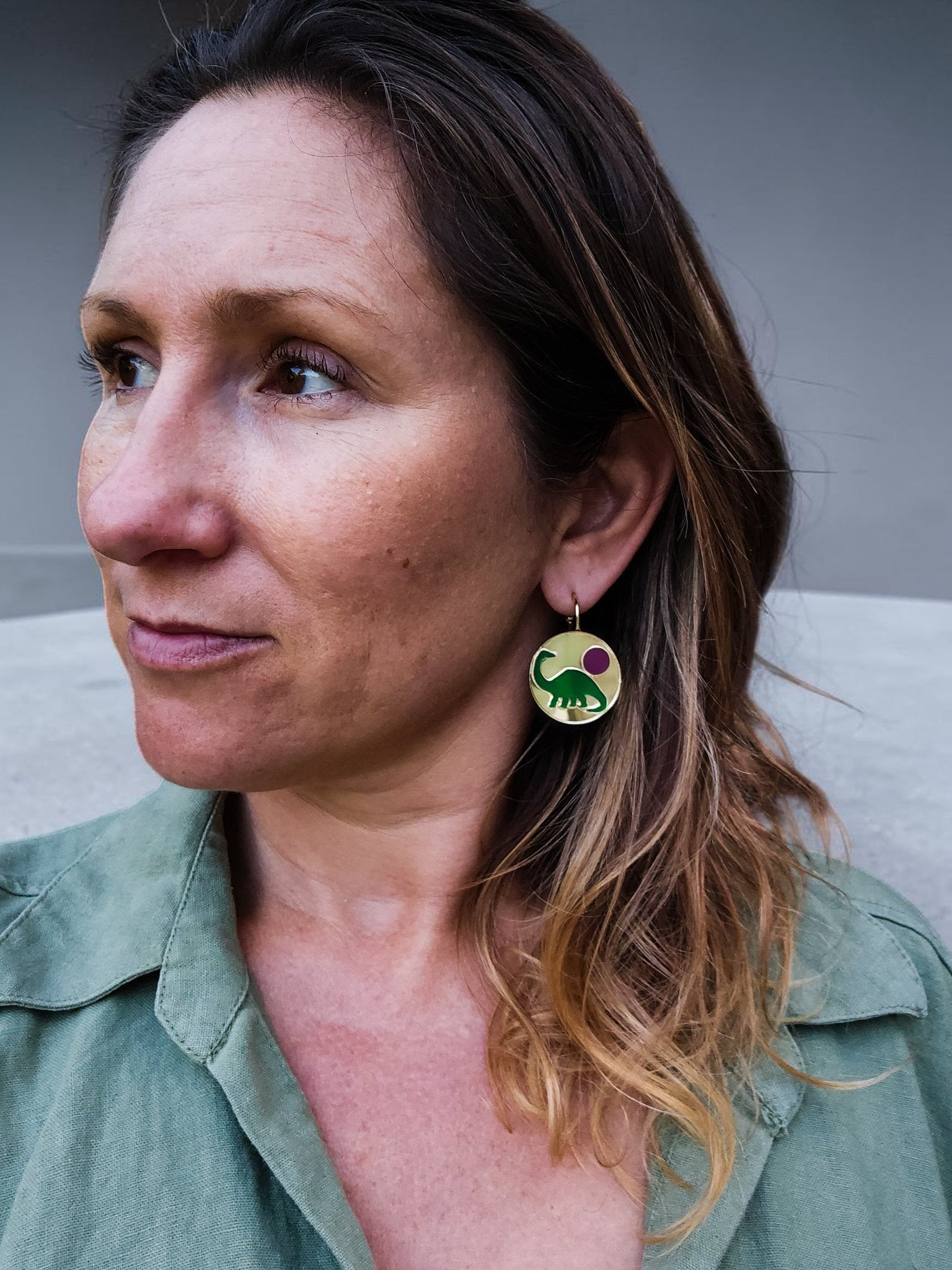 Morti's Fun Club Tiny Dino earrings featuring a green dinosaur and pink moon on gold acrylic hoops worn by a woman.