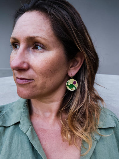 Morti's Fun Club Tiny Dino earrings featuring a green dinosaur and pink moon on gold acrylic hoops worn by a woman.